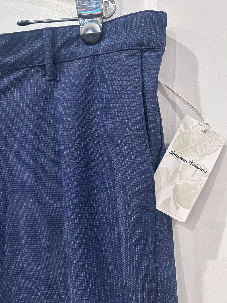 May include: Close-up of navy blue pants with a textured pattern. The pants have a pocket and a tag that reads "Tommy Bahama". The pants are hanging on a hanger.