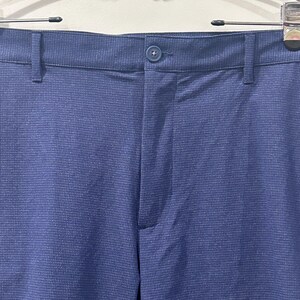 May include: Close-up of blue pants with a textured weave. The pants have a button closure and belt loops. The fabric appears to be a solid blue color. The pants are hanging on a metal hanger.