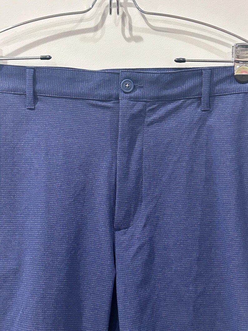 May include: Close-up of blue pants with a textured weave. The pants have a button closure and belt loops. The fabric appears to be a solid blue color. The pants are hanging on a metal hanger.