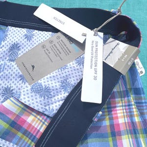 K&ouml;nnte beinhalten: Nahaufnahme einer mehrfarbigen karierten Shorts mit marineblauem Bund. Die Shorts haben ein wei&szlig;es Ananas-Print-Futter. Angebrachte Etiketten lauten "STRETCH", "SONNENSCHUTZ UPF 30" und "ISLAND ZONE".