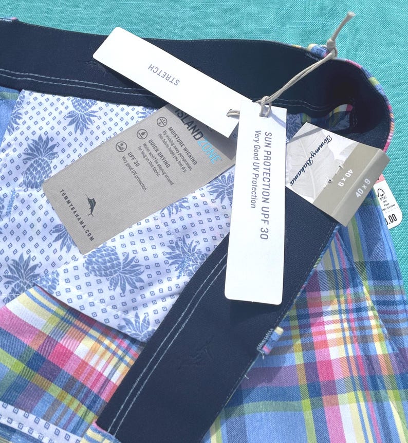 K&ouml;nnte beinhalten: Nahaufnahme einer mehrfarbigen karierten Shorts mit marineblauem Bund. Die Shorts haben ein wei&szlig;es Ananas-Print-Futter. Angebrachte Etiketten lauten "STRETCH", "SONNENSCHUTZ UPF 30" und "ISLAND ZONE".