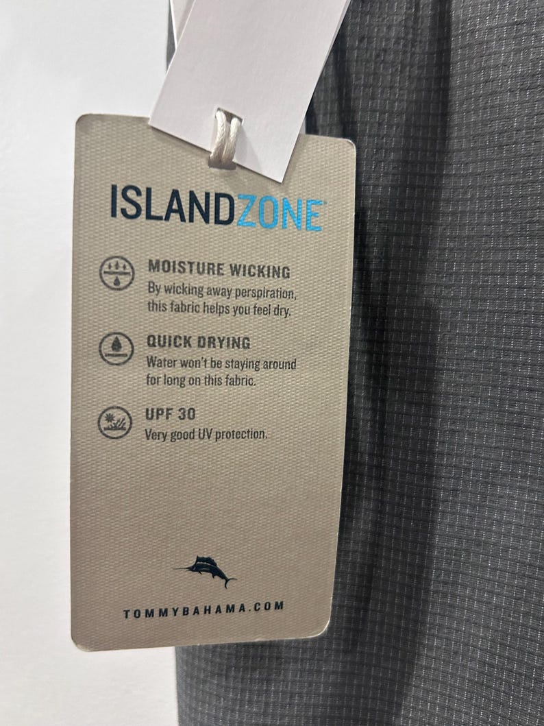 May include: A gray fabric tag with the text "ISLANDZONE" in blue. The tag details moisture-wicking, quick-drying, and UPF 30 UV protection properties. The Tommy Bahama logo and website are at the bottom. The fabric is a textured gray.