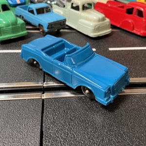 May include: A blue convertible toy car with a detailed front grille and open top. Several other toy vehicles, including a green van, a light blue pickup truck, and a red truck, are visible in the background. The toys are on a black surface.
