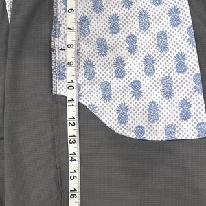May include: Close-up of gray shorts with a patterned interior featuring blue pineapples on a white background. The shorts have a gray waistband with a dolphin design and a tag that reads "Tommy Bahama". The shorts measure approximately 22 inches in length.