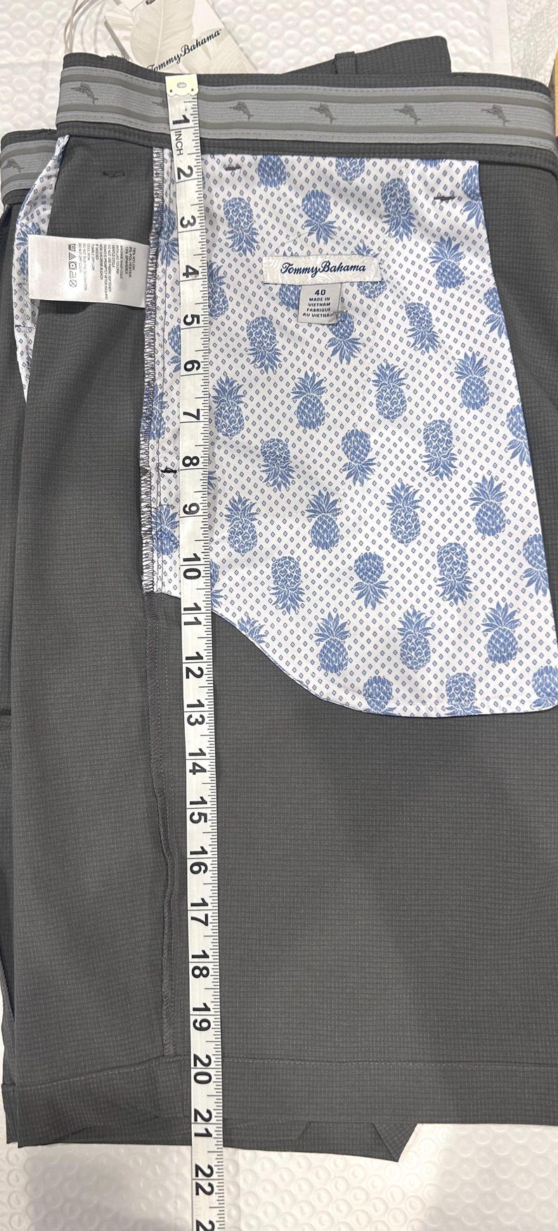May include: Close-up of gray shorts with a patterned interior featuring blue pineapples on a white background. The shorts have a gray waistband with a dolphin design and a tag that reads "Tommy Bahama". The shorts measure approximately 22 inches in length.