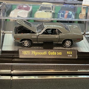 May include: A gray 1971 Plymouth Cuda 340 model car with its hood open, displayed in a clear plastic case. The model car has black tires and a black interior. The text "1971 Plymouth Cuda 340" is visible on a small plaque.