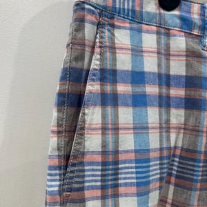 Peut inclure: Gros plan sur un short &agrave; carreaux bleu, blanc, rose et gris. Le short a une poche avant et une fermeture boutonn&eacute;e. Le tissu a une texture tiss&eacute;e.