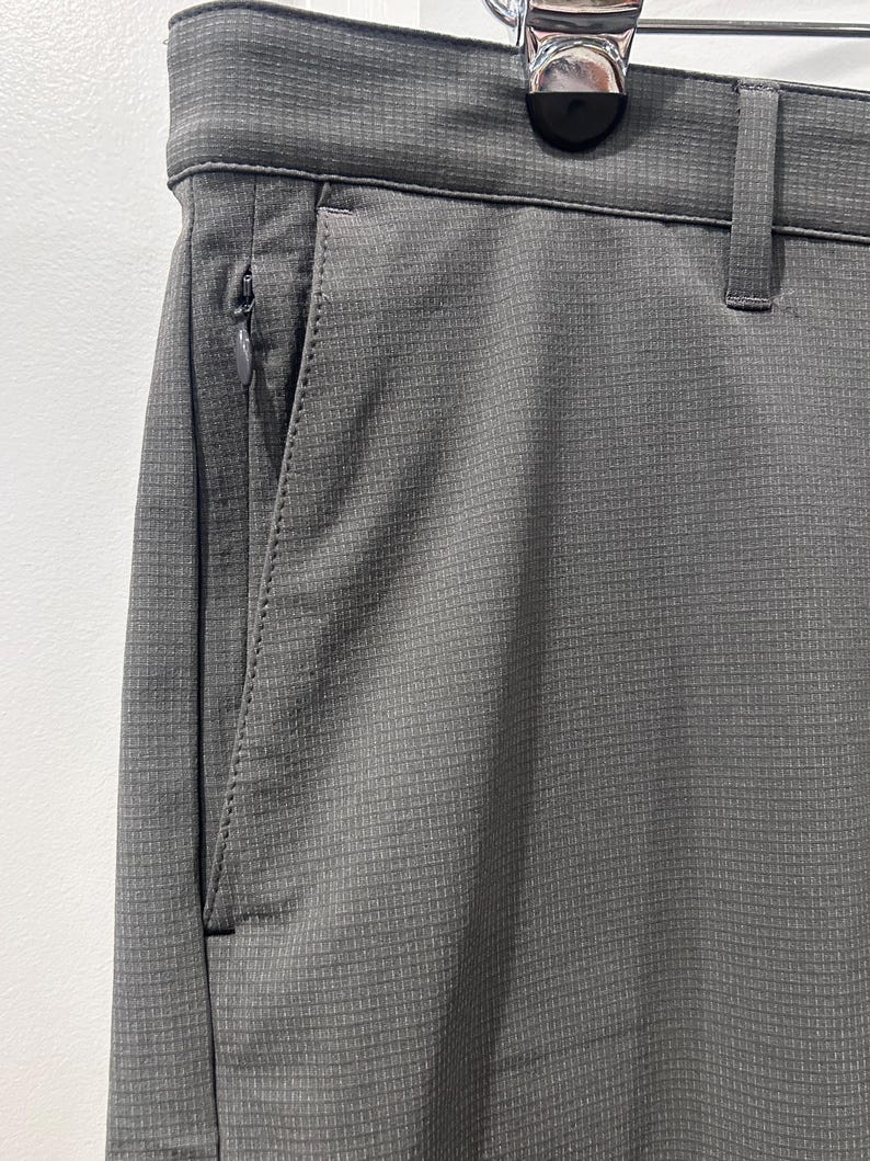 May include: Close-up of gray pants with a textured, grid-like pattern. The pants have a zippered pocket on the side and a belt loop. The fabric appears to be a lightweight, breathable material, suitable for warm weather.