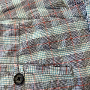 May include: Close-up of a pair of shorts with a plaid pattern in shades of blue, red, and white. The shorts have a button closure and a pocket detail. The fabric appears to be cotton or a similar material.