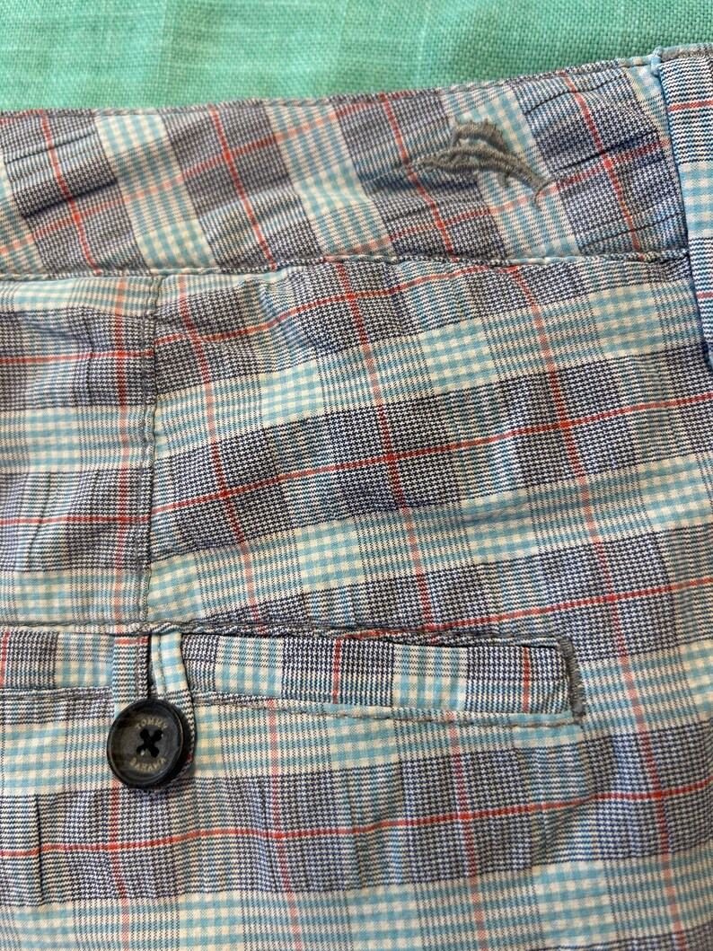 May include: Close-up of a pair of shorts with a plaid pattern in shades of blue, red, and white. The shorts have a button closure and a pocket detail. The fabric appears to be cotton or a similar material.
