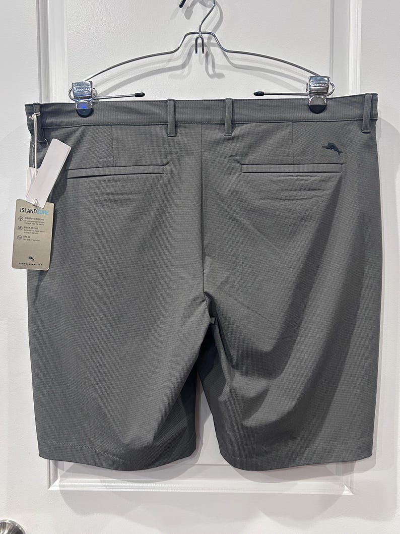 May include: Gray golf shorts with a textured pattern, featuring back pockets and belt loops. A tag with the brand name "ISLAND" is attached. The shorts are hanging on a metal hanger.