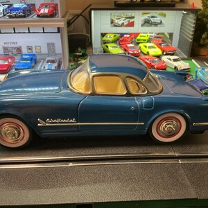 May include: A blue die-cast model car with a hardtop, white-walled tires, and red accents. The model car has a gold-colored windshield frame and the word "Chevrolet" is written on the side. The car is on a black track.