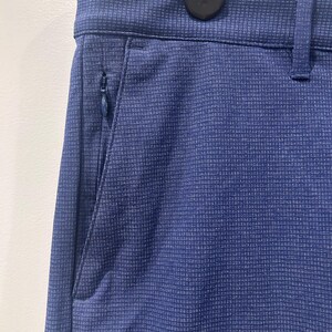 May include: Close-up of navy blue pants with a subtle grid pattern. The pants feature a zippered pocket on the side and a belt loop. The fabric has a textured appearance, and the pants are hanging on a hanger.