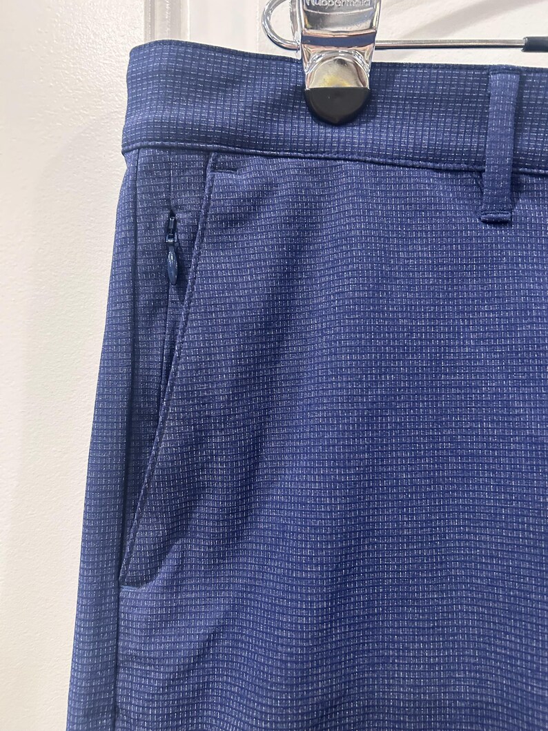 May include: Close-up of navy blue pants with a subtle grid pattern. The pants feature a zippered pocket on the side and a belt loop. The fabric has a textured appearance, and the pants are hanging on a hanger.