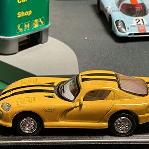 May include: A yellow Dodge Viper toy car with black racing stripes. The miniature car has detailed silver wheels and a sleek design. A blue and white race car is in the background, along with a green sign that says "Car Shop".
