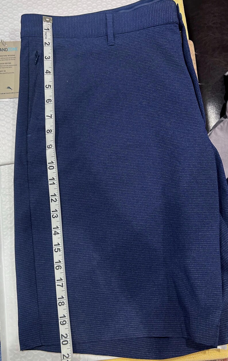 May include: Navy blue shorts with a textured pattern. The shorts are measured with a ruler, showing a length of approximately 20 inches. The shorts have a button and a zipper closure.