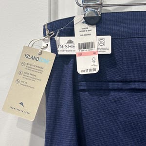 May include: Navy blue men's shorts with a subtle grid pattern, hanging from a metal clip. The shorts have a tag that reads "Island Zone" and "Moisture Wicking". The shorts are size 40.