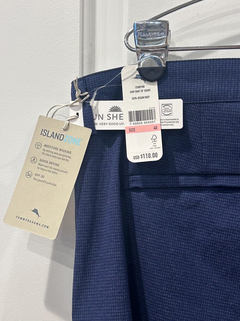 May include: Navy blue men's shorts with a subtle grid pattern, hanging from a metal clip. The shorts have a tag that reads "Island Zone" and "Moisture Wicking". The shorts are size 40.