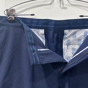 May include: Close-up of navy blue dress pants hanging on a silver hanger. The pants are made of a textured fabric and feature a zipper and button closure. The interior lining has a white and blue pineapple pattern.