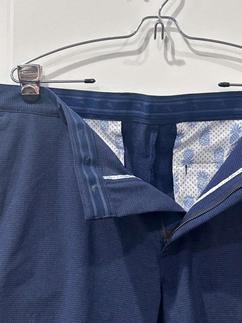 May include: Close-up of navy blue dress pants hanging on a silver hanger. The pants are made of a textured fabric and feature a zipper and button closure. The interior lining has a white and blue pineapple pattern.
