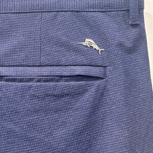May include: Close-up of blue dress pants with a subtle textured pattern. A silver-colored fish emblem is affixed near the back pocket. The pants are hanging on a hanger.
