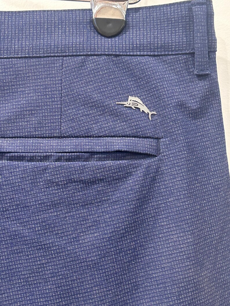 May include: Close-up of blue dress pants with a subtle textured pattern. A silver-colored fish emblem is affixed near the back pocket. The pants are hanging on a hanger.