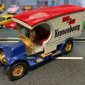 May include: A vintage-style toy truck with a red roof and blue body. The truck features gold-colored wheels and trim, and the side displays the "Kronenbourg INTERNATIONAL BEER" logo. The truck is parked on a gray surface with other toy cars in the background.