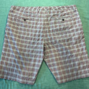 May include: A pair of knee-length shorts with a plaid pattern in shades of pink, white, and gray. The shorts have two back pockets and a button closure. The fabric appears to be lightweight.