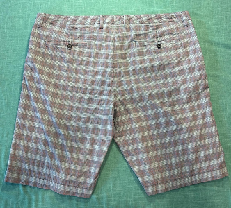 May include: A pair of knee-length shorts with a plaid pattern in shades of pink, white, and gray. The shorts have two back pockets and a button closure. The fabric appears to be lightweight.