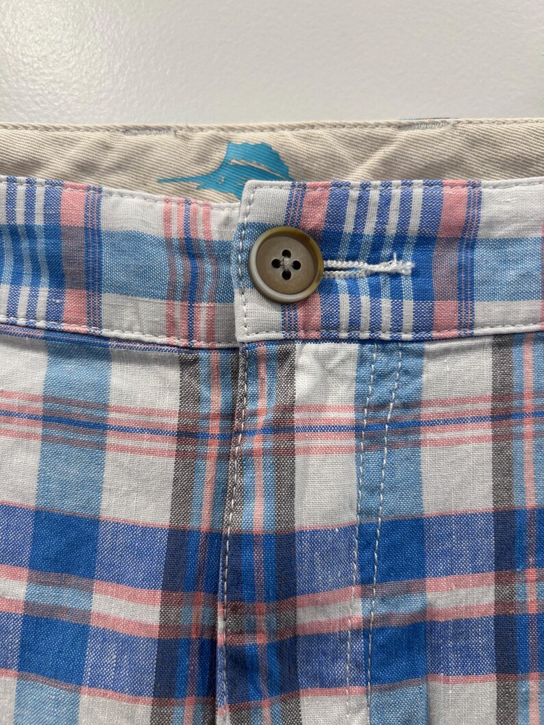 Peut inclure: Gros plan sur un short &agrave; carreaux bleu, rose et blanc. Un bouton marron ferme la ceinture. Le tissu semble l&eacute;ger. Le motif &agrave; carreaux est un motif classique.