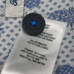 May include: Close-up of a Tommy Bahama shirt tag and button. The gray button has the brand name and a blue cross-stitch design. The shirt fabric is white with a blue pineapple pattern. The tag includes the brand name and address.