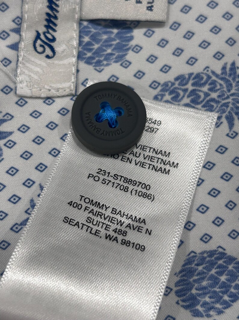 May include: Close-up of a Tommy Bahama shirt tag and button. The gray button has the brand name and a blue cross-stitch design. The shirt fabric is white with a blue pineapple pattern. The tag includes the brand name and address.