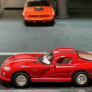 May include: A red toy car with silver racing stripes and silver wheels. The toy car has the words "Viper" and "SRT" on the side. An orange toy car is in the background.