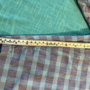 May include: A blue, red, and white plaid shirt sleeve is measured with a yellow and white measuring tape. The tape shows the sleeve is approximately 10 inches wide. The shirt is on a turquoise background.