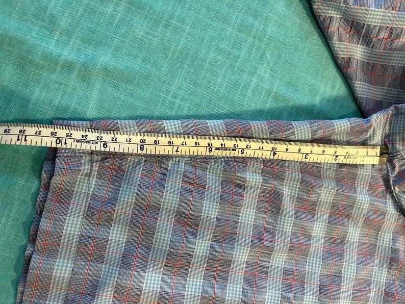 May include: A blue, red, and white plaid shirt sleeve is measured with a yellow and white measuring tape. The tape shows the sleeve is approximately 10 inches wide. The shirt is on a turquoise background.