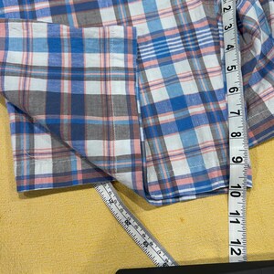 Peut inclure: Une chemise &agrave; carreaux bleue, blanche et rose avec une manche et une poche visibles. La chemise est mesur&eacute;e avec une r&egrave;gle, montrant une longueur d'environ 30 cm.