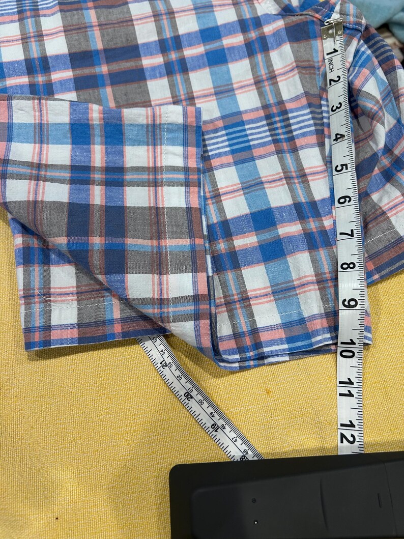Peut inclure: Une chemise &agrave; carreaux bleue, blanche et rose avec une manche et une poche visibles. La chemise est mesur&eacute;e avec une r&egrave;gle, montrant une longueur d'environ 30 cm.