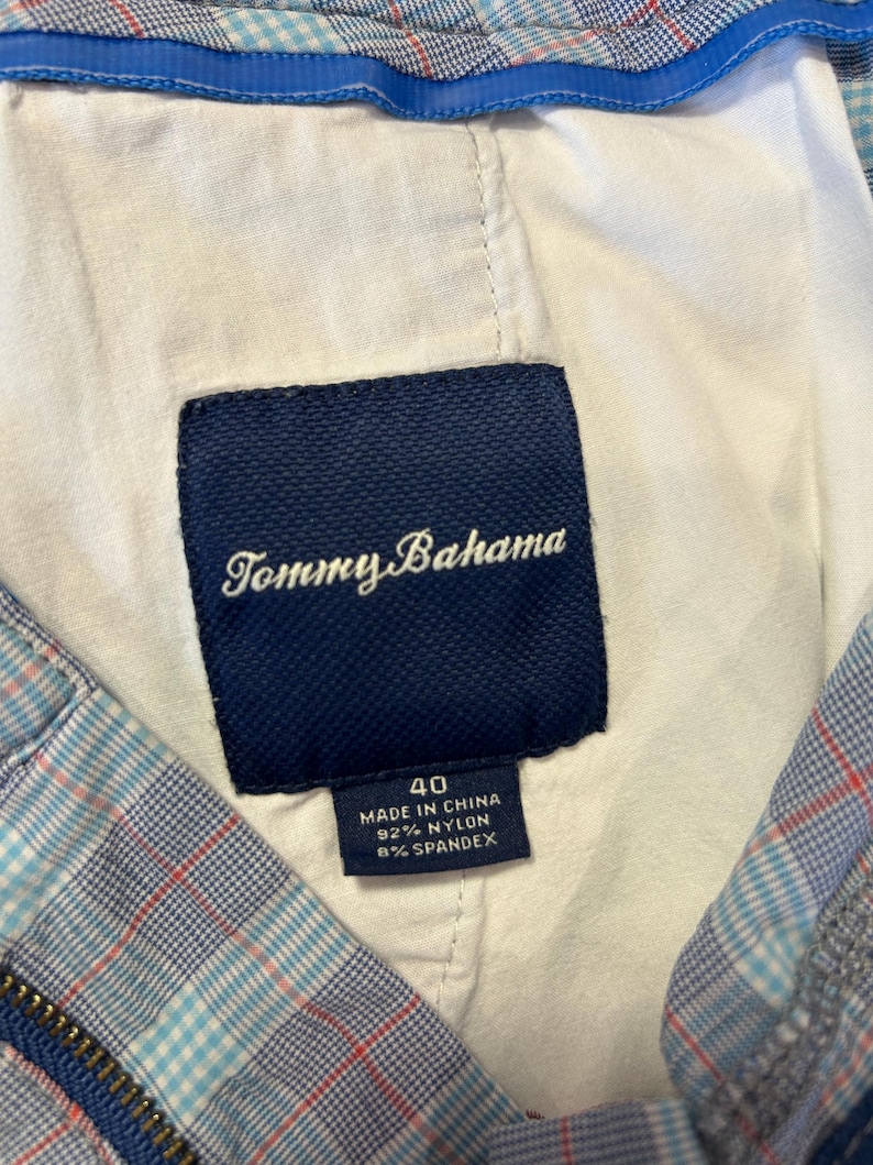 May include: Close-up of a navy blue Tommy Bahama label, size 40, sewn into the lining of a plaid garment. The label indicates the garment is made in China of 92% nylon and 8% spandex. The plaid pattern includes blue, red, and white.