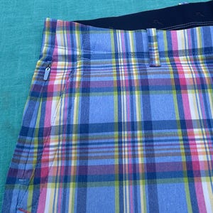 K&ouml;nnte beinhalten: Nahaufnahme einer karierten Shorts in Blau-, Rosa-, Gelb- und Wei&szlig;t&ouml;nen. Die Shorts haben einen schwarzen Bund und eine Rei&szlig;verschlusstasche. Das Karomuster ist ein klassisches Design.