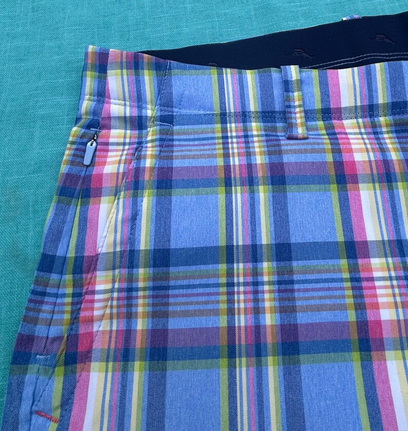 K&ouml;nnte beinhalten: Nahaufnahme einer karierten Shorts in Blau-, Rosa-, Gelb- und Wei&szlig;t&ouml;nen. Die Shorts haben einen schwarzen Bund und eine Rei&szlig;verschlusstasche. Das Karomuster ist ein klassisches Design.