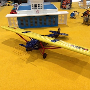 May include: A yellow and blue toy airplane with red accents. The airplane has a propeller and wheels. The airplane is sitting on a yellow surface. The text "Airship The Amigo" is printed on the side of the airplane.