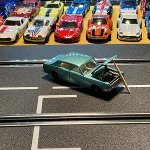 May include: A teal-colored toy car with its hood open, propped up by a wooden stick, sits on a black surface with white lines. A collection of colorful toy race cars are lined up in the background.