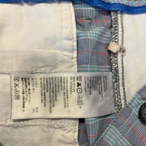 May include: Close-up of a pair of plaid shorts with a blue, red, and white pattern. The shorts have a white interior lining and a blue waistband. A tag with washing instructions is visible.