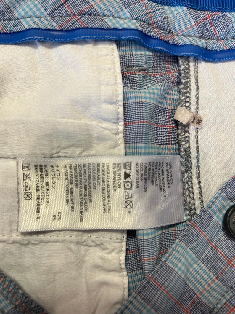 May include: Close-up of a pair of plaid shorts with a blue, red, and white pattern. The shorts have a white interior lining and a blue waistband. A tag with washing instructions is visible.