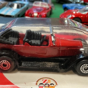 May include: A red vintage toy car with black fenders and wheels, encased in clear plastic. The car has a detailed interior and is part of a collection of toy cars. The wheels have a silver rim.