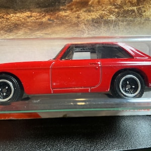 May include: A bright red die-cast toy car in a clear plastic blister pack. The vehicle features a two-door coupe body style, black wheels with silver rims, and a visible interior. The side profile shows a long hood and a sloped rear window.