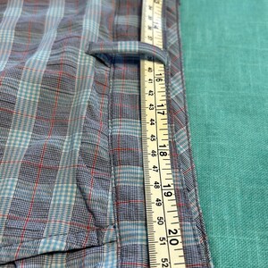 May include: Close-up of a blue, red, and white plaid shirt with a measuring tape. The tape shows measurements up to 55 inches. The shirt has a button and a pocket.