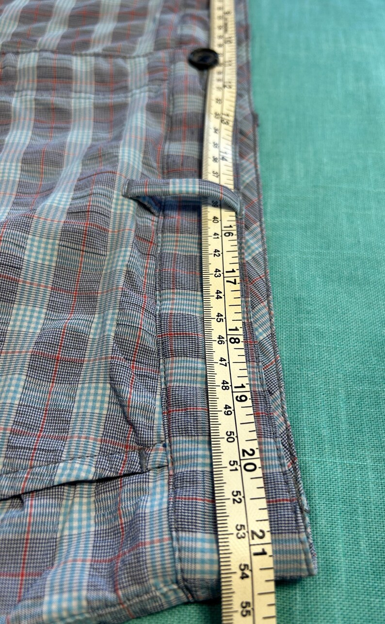 May include: Close-up of a blue, red, and white plaid shirt with a measuring tape. The tape shows measurements up to 55 inches. The shirt has a button and a pocket.