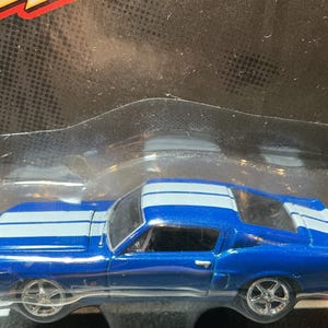 May include: A blue die-cast Shelby GT500KR toy car featuring dual white racing stripes running along the hood, roof, and trunk. The vehicle has silver-colored wheels and is encased in clear plastic packaging with black and red cardboard backing.