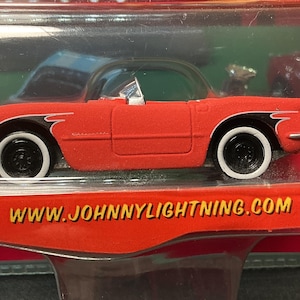 May include: A red toy car with black and white wheels and a black roof. The car has white accents and the text "CORVETTE" is visible. The website address "WWW.JOHNNYLIGHTNING.COM" is printed on the packaging.